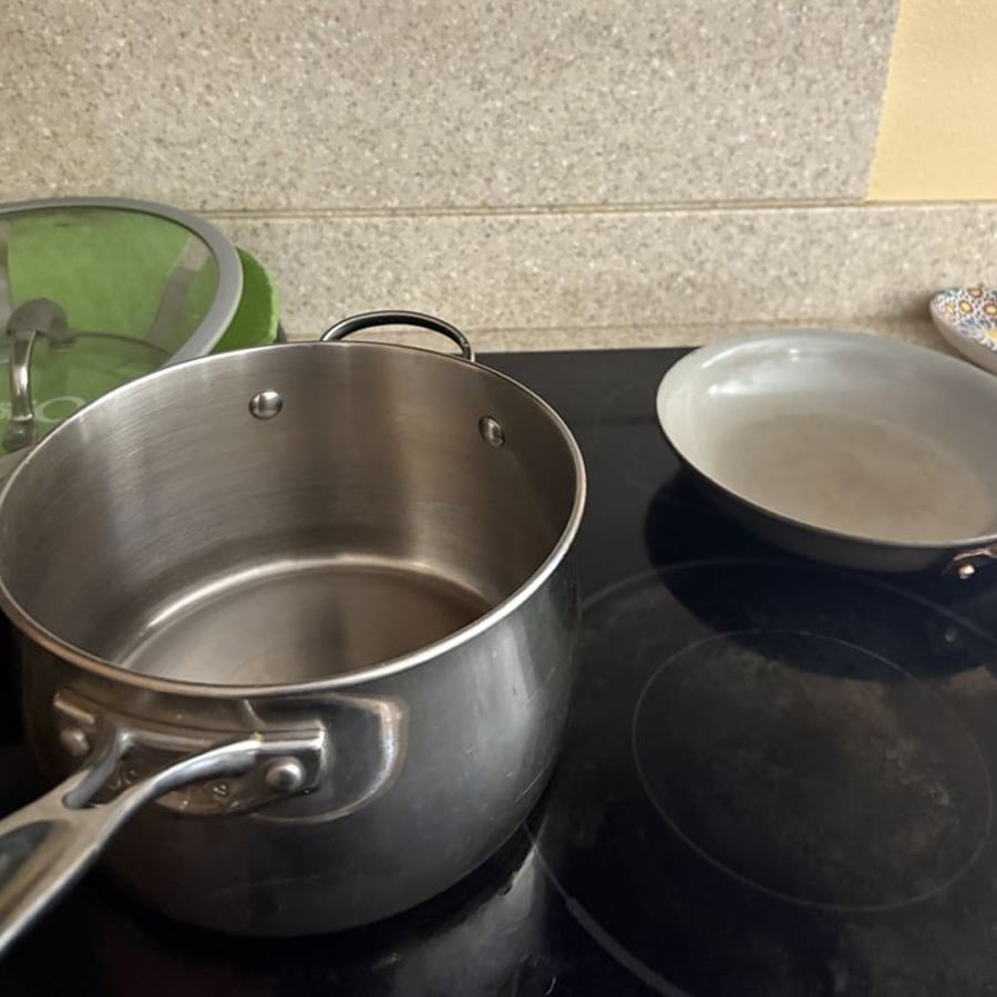 A stainless steel pot and a metal frying pan sit on a black stove next to a beige countertop. A green dish rack with a glass lid is visible in the background.