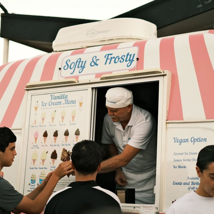 A man in a white uniform serves ice cream from a pink and white striped truck labeled "Softy & Frosty" to two customers standing at the window. The menu displays various ice cream options and a vegan option.