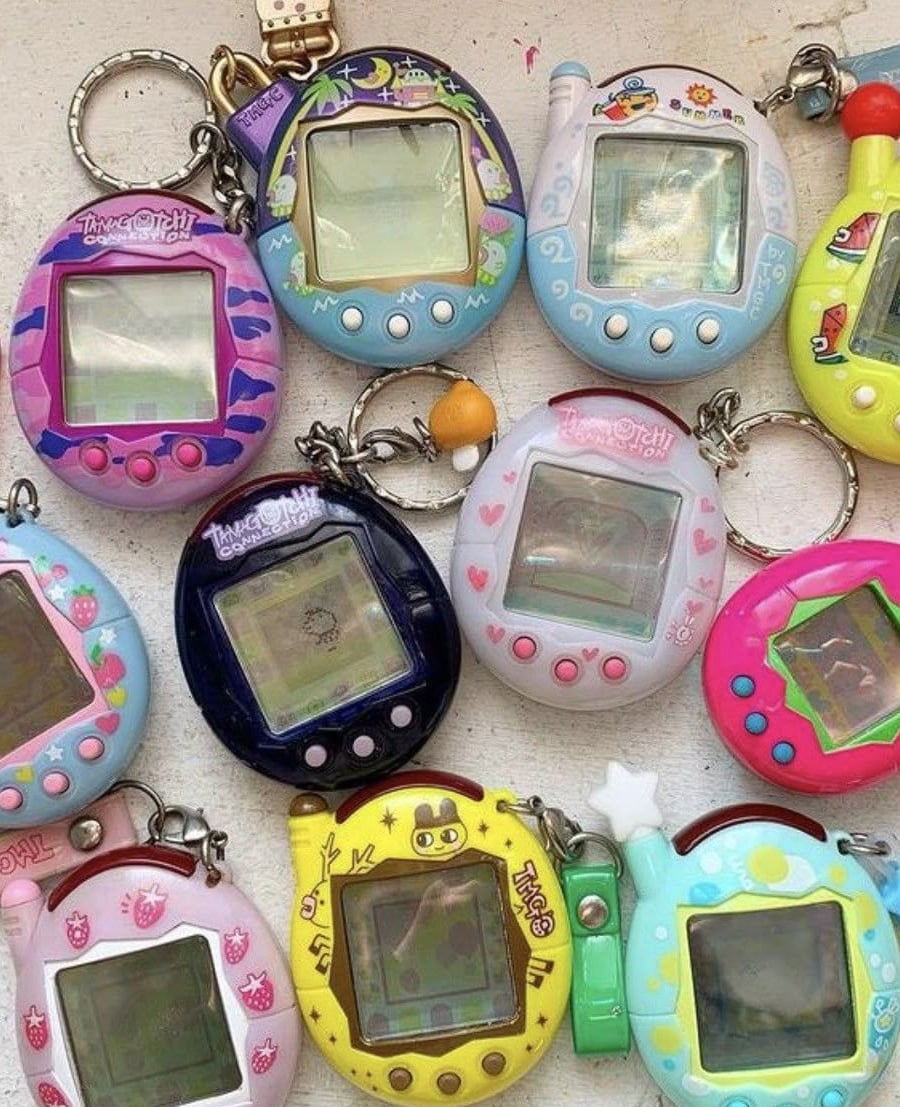 A collection of colorful Tamagotchi virtual pet keychains arranged on a flat surface. Each device has a small screen and distinctive designs, including patterns, stars, and cartoon faces.