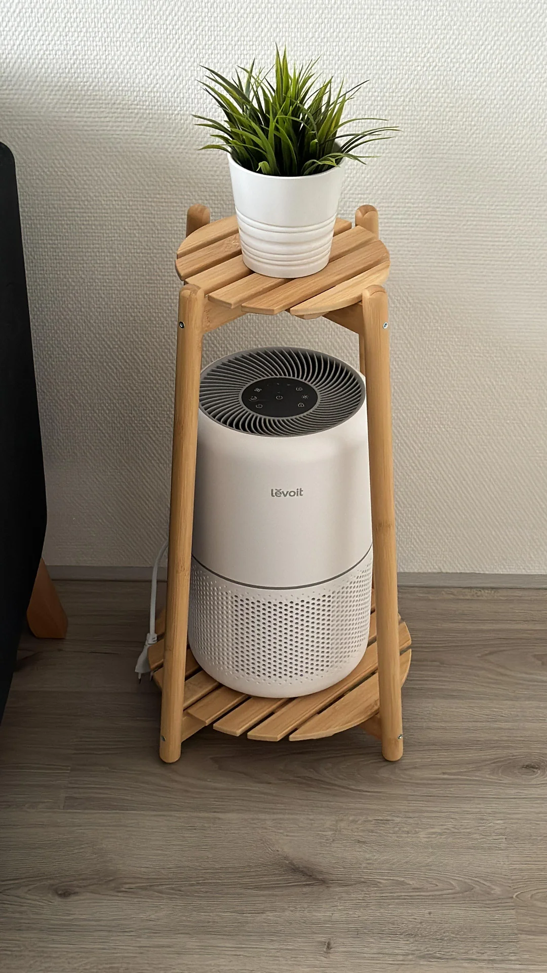 A small white air purifier sits on the bottom shelf of a wooden stand, with a green potted plant placed on the top shelf. The stand is on a light wood floor against a textured white wall.