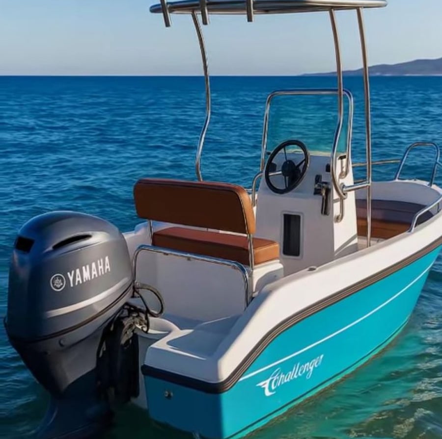 A small turquoise and white motorboat with a Yamaha outboard engine floats on clear blue water. The boat has tan seats, a steering wheel, and a metal canopy. "Challenger" is written on the side. Distant land is visible.