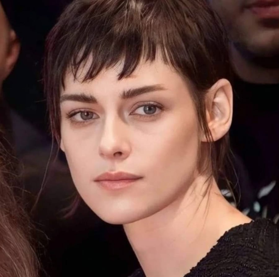 A woman with short dark hair and bangs looks slightly to the side with a neutral expression. She is wearing a black top and appears to be in an indoor setting with others in the background.
