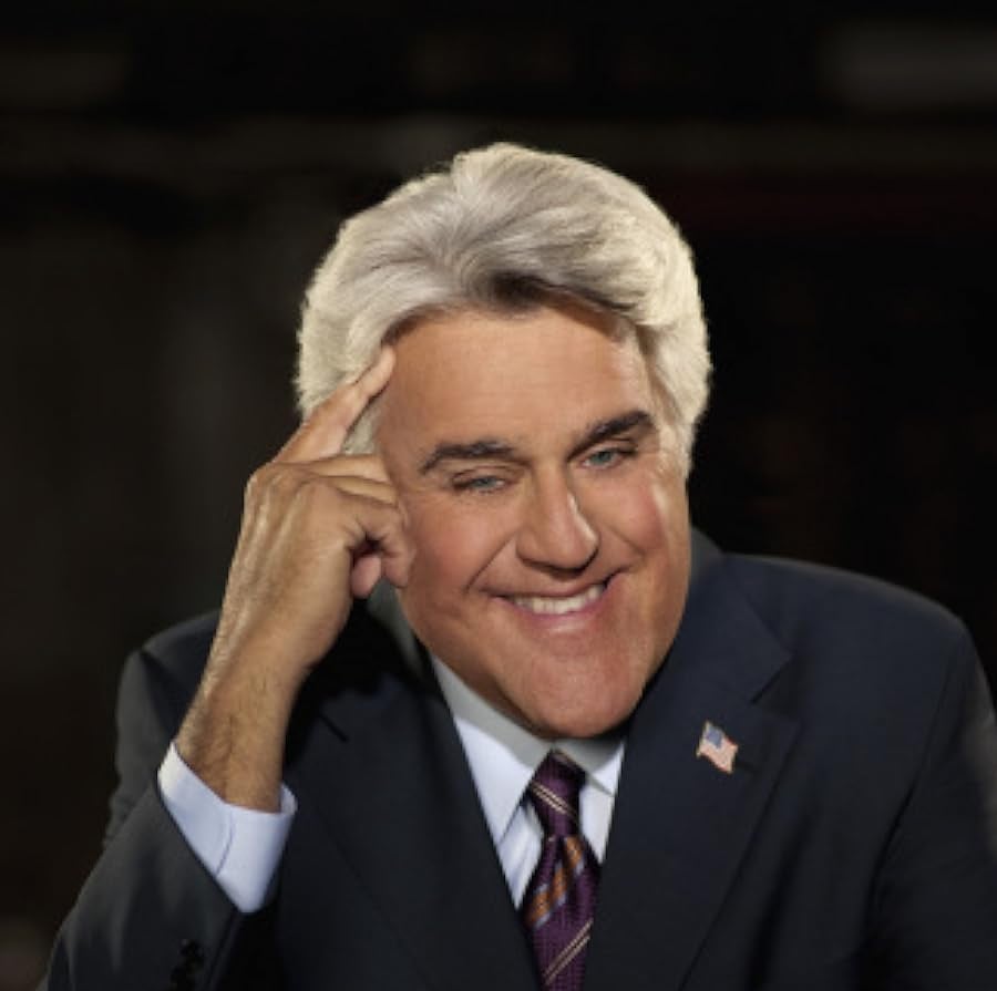 A man with gray hair wearing a dark suit, striped tie, and an American flag pin sits smiling with one hand touching the side of his head against a dark background.