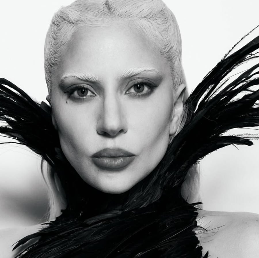 A person with platinum blond hair and strong facial features stares seriously at the camera, wearing a dramatic black feathered collar against a plain background. The photo is in black and white.