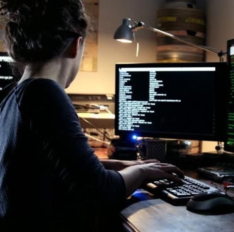 A person with dark hair sits at a desk, typing on a keyboard and looking at a computer monitor displaying lines of code or text in a dimly lit room with various equipment and monitors.