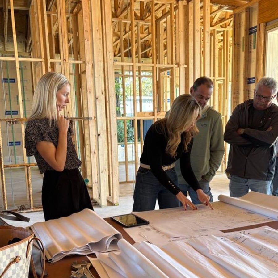 Four people stand in a partially constructed building, examining architectural blueprints on a table covered with plans and documents. Exposed wooden framing surrounds them as they discuss construction details.