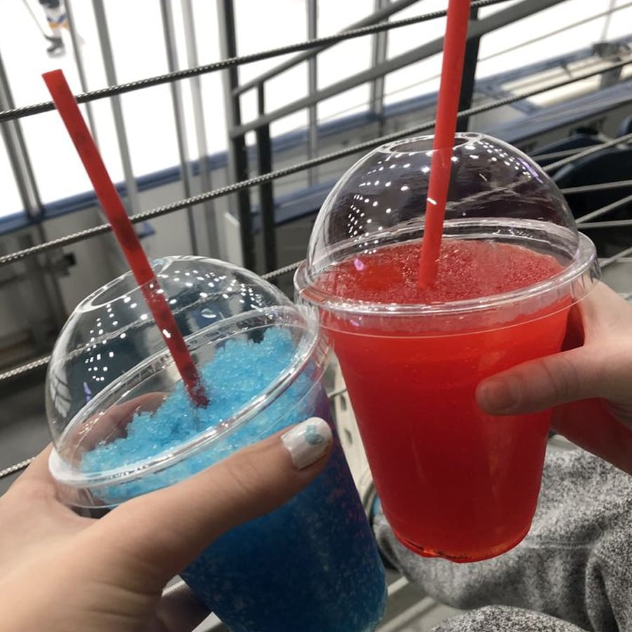 Two hands holding plastic cups with dome lids and red straws; one cup contains a blue slushie, the other a red slushie. Metal railings and seating are visible in the background, suggesting a public venue.