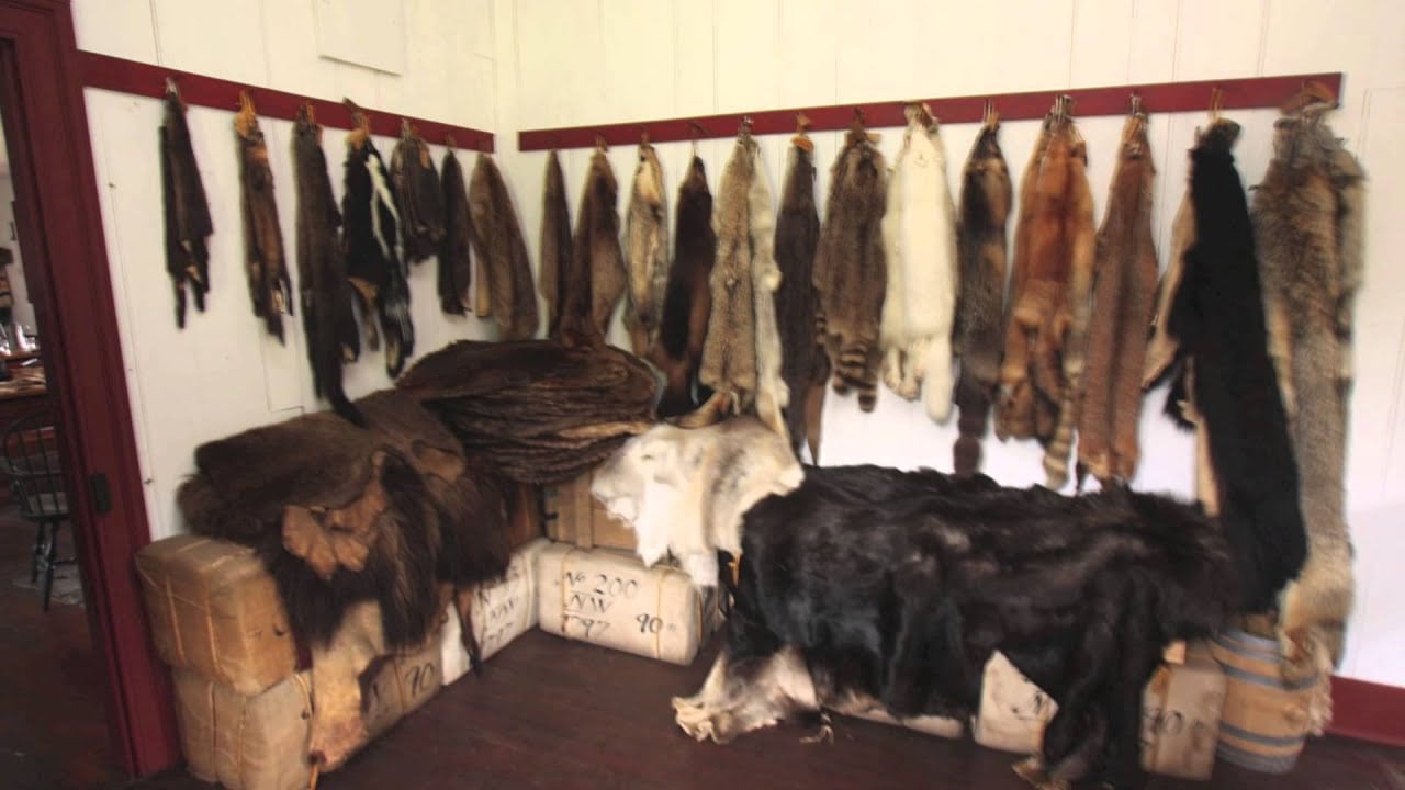 Animal pelts and furs hang on hooks along two walls in a corner room, with several large, bundled furs and animal hides stacked on top of burlap sacks on the wooden floor below.