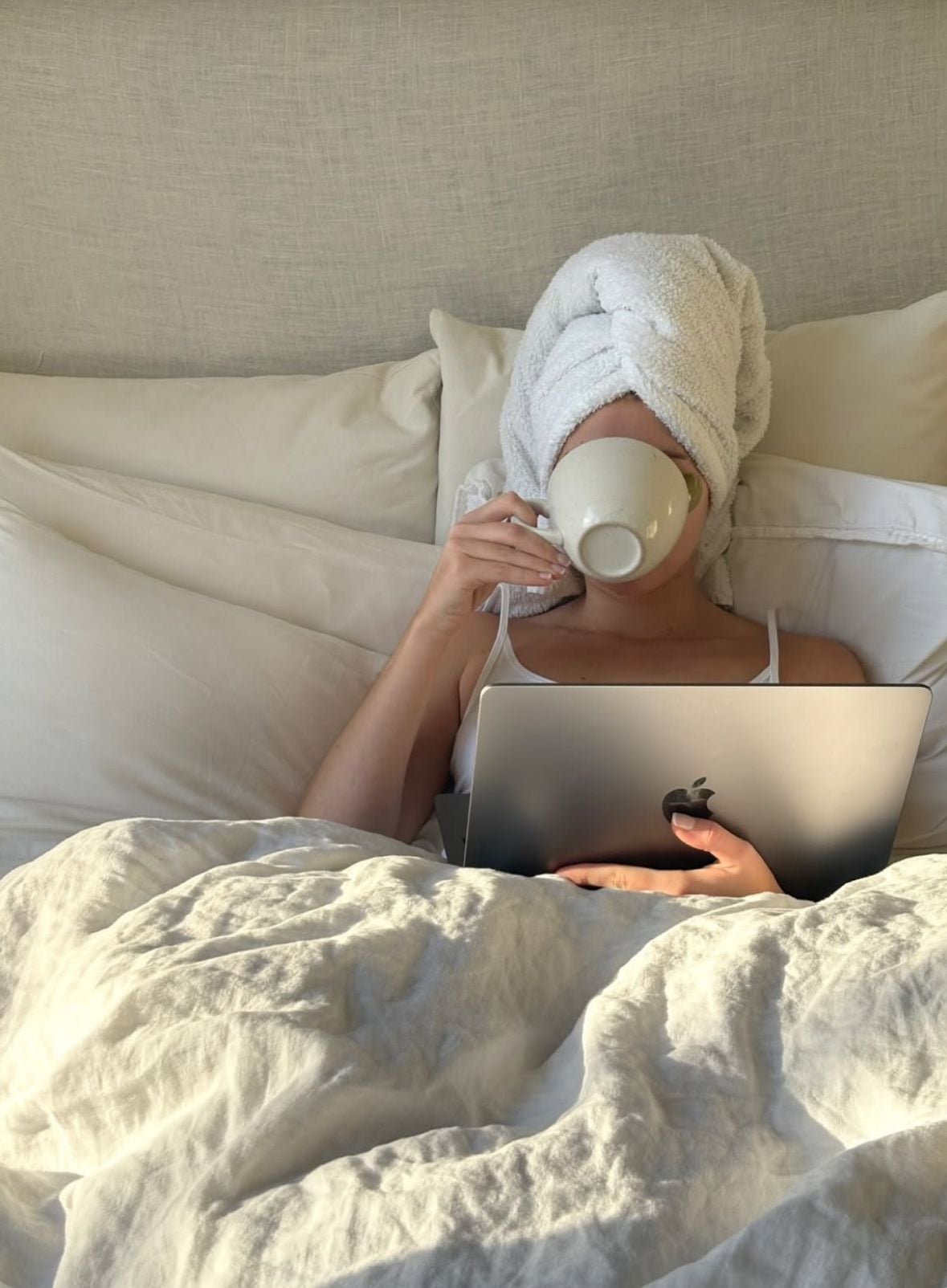 A person sitting up in bed with a towel wrapped around their head, sipping from a mug, and using an Apple laptop, surrounded by white bedding and pillows.
