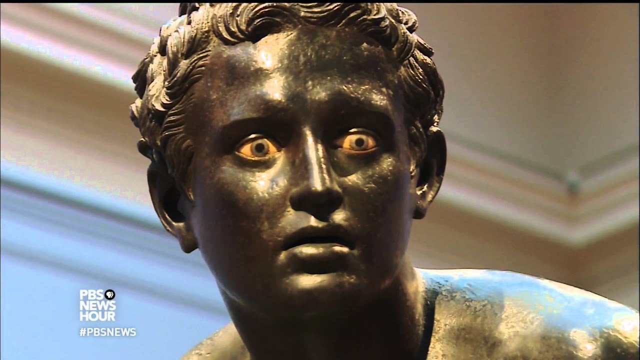 Close-up of an ancient bronze statue with detailed curly hair and striking, inlaid eyes, giving it a lifelike and intense expression. PBS NewsHour and PBSNews text logos are visible in the lower left corner.