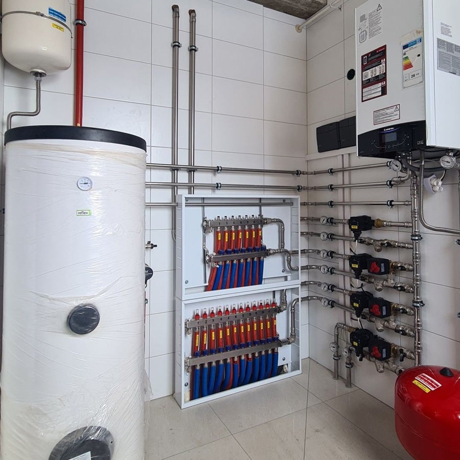 A utility room with a white boiler tank, manifold with red and blue pipes, control panel, wall-mounted boiler, gauges, and various metal pipes against white tiled walls.