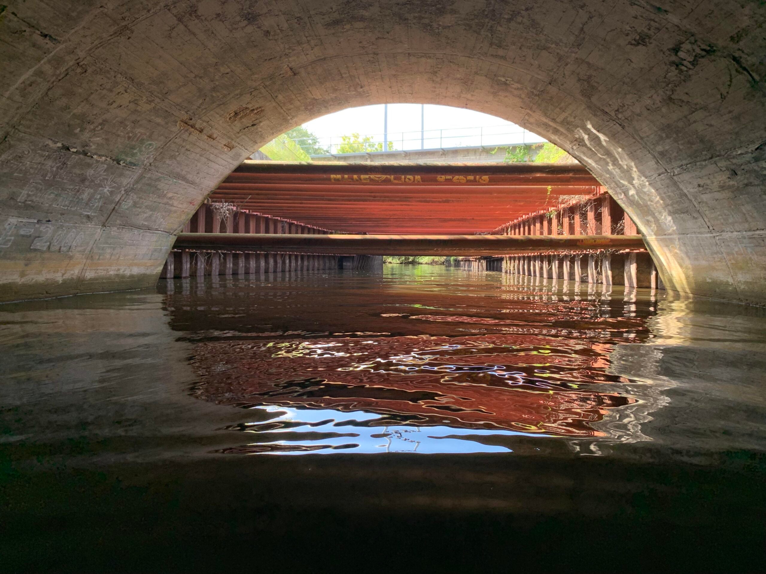 View from under a concrete tunnel with water below, looking out toward sunlight and a metal bridge structure with reflections in the water. Trees and a blue sky are visible beyond the bridge.