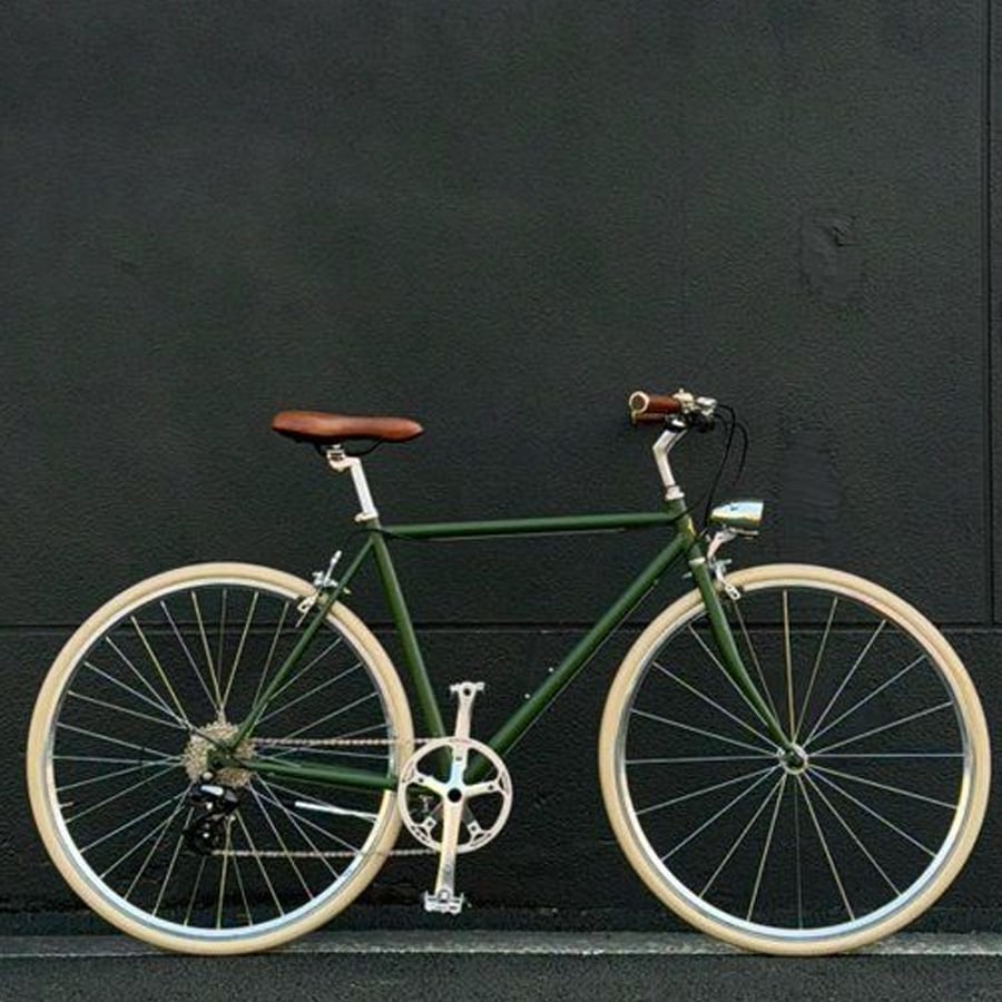 A green single-speed bicycle with tan tires and a brown leather seat is parked in front of a plain dark gray wall.