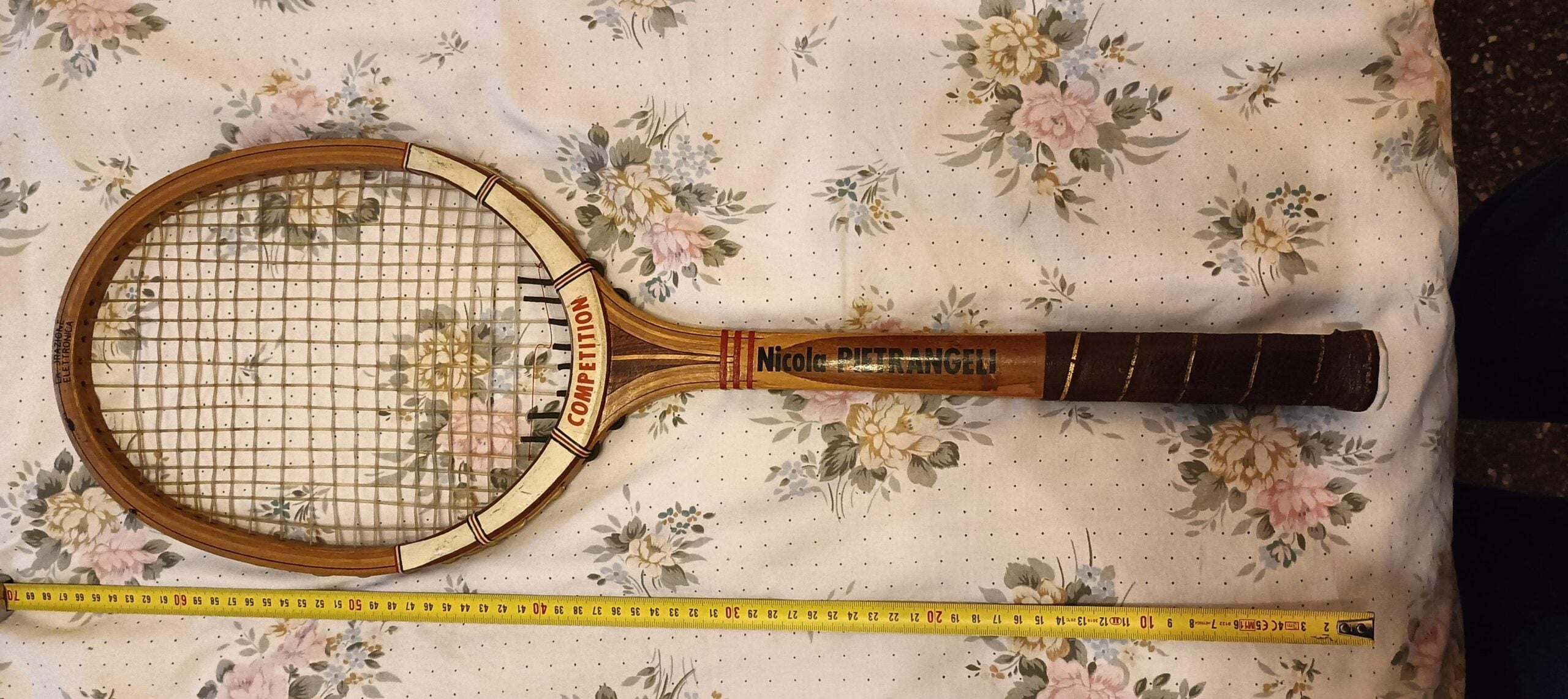 A vintage wooden tennis racket with "CONTROL" written on the frame, "Nicol" and "CHAMPION" on the handle, lies on a floral-patterned fabric next to a yellow measuring tape.
