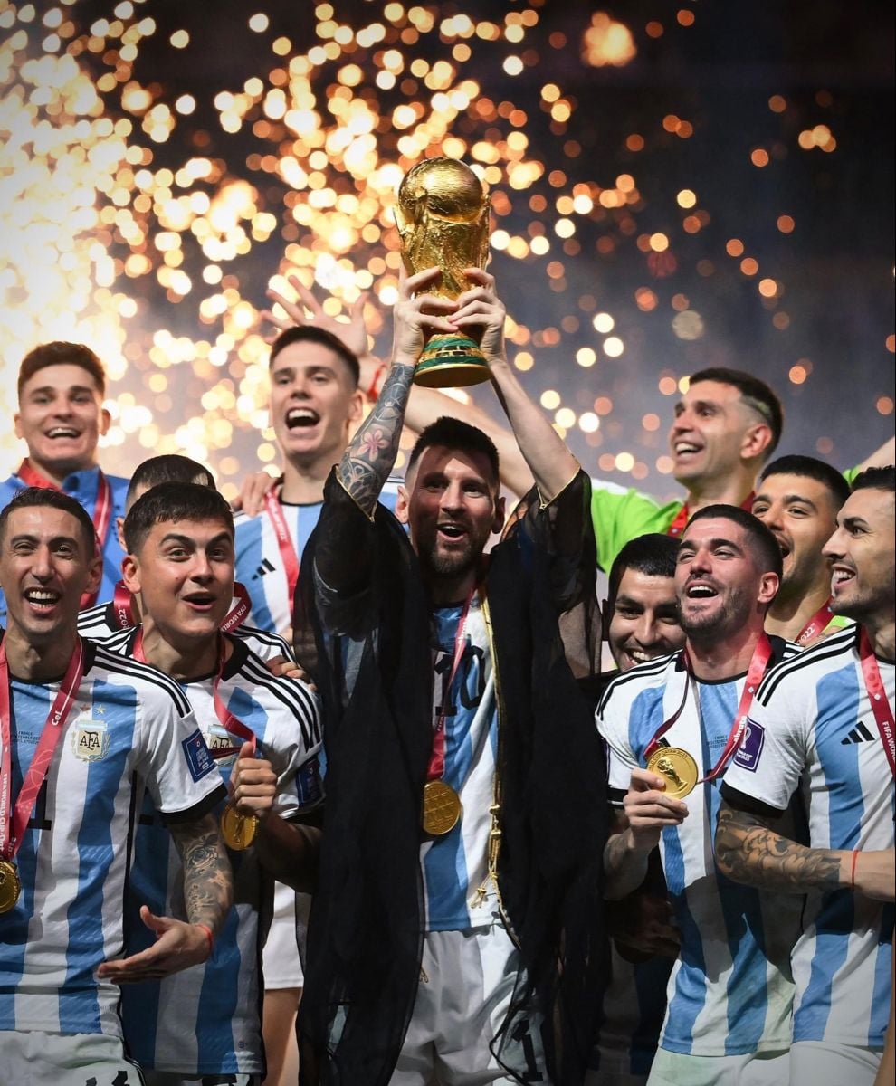 A soccer player in an Argentina jersey lifts a gold trophy above his head, surrounded by jubilant teammates wearing medals, with fireworks sparkling in the background.