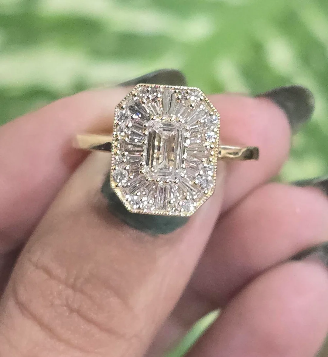 A hand holding a gold ring with a large, rectangular center stone surrounded by smaller baguette and round diamonds, set against a blurred green background.