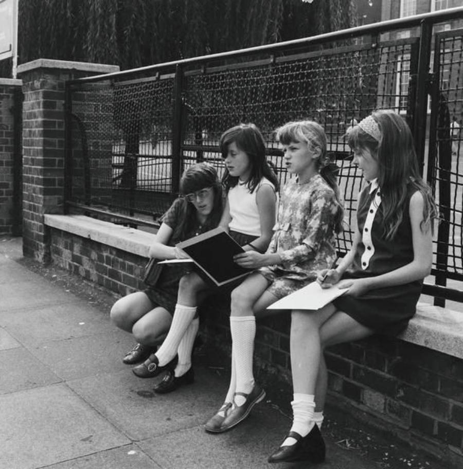 Four girls sit on a low brick wall with notebooks in their laps, appearing to draw or write. They wear dresses and knee-high socks, and a black iron fence runs behind them along the sidewalk.