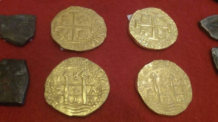 Four gold coins with intricate, worn designs are displayed on a red surface, with parts of two dark, irregularly shaped objects visible near the edges. The coins feature cross and crest patterns.