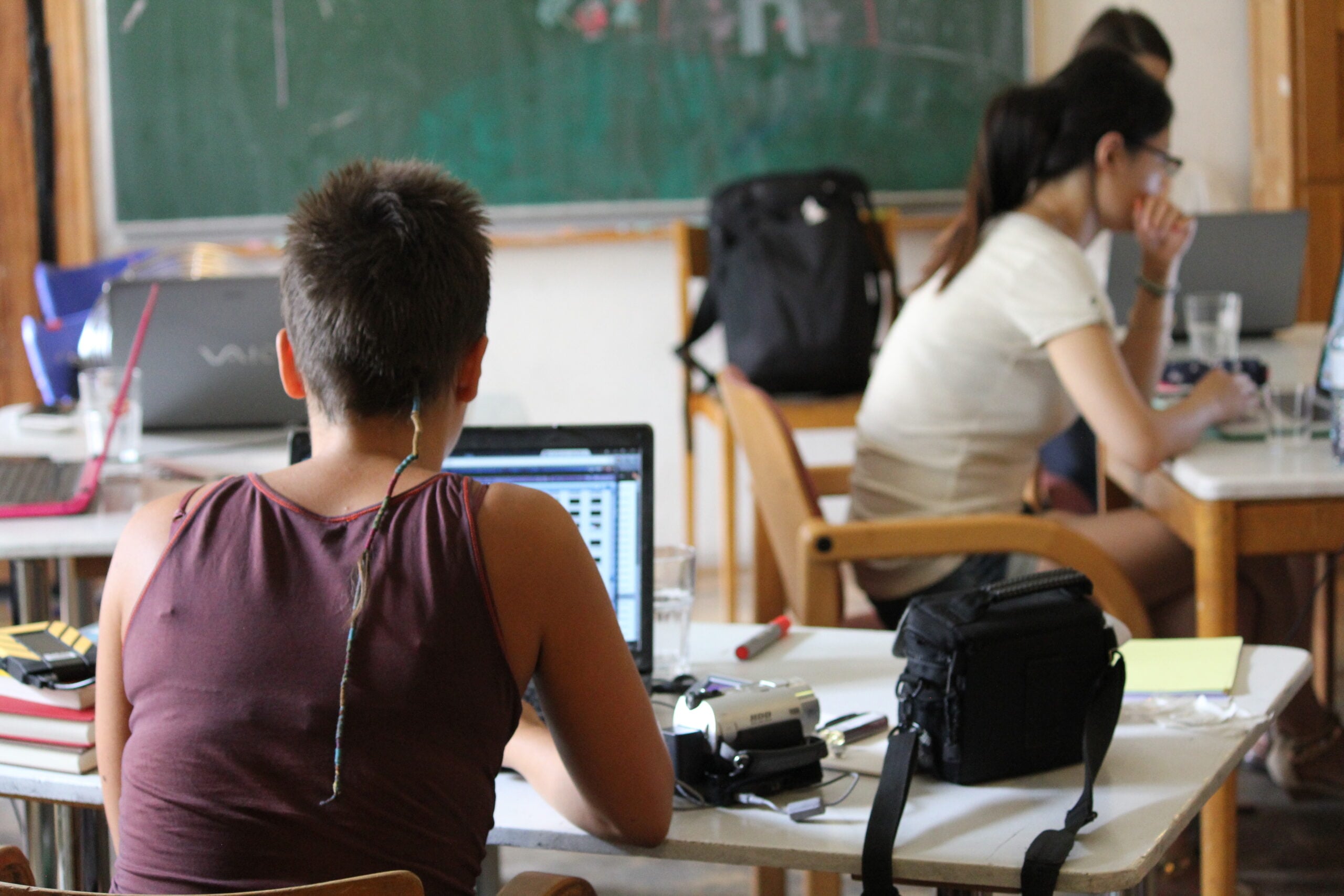 Several people sit at tables working on laptops in a classroom setting, with a chalkboard in the background and various electronic devices and bags on the tables.