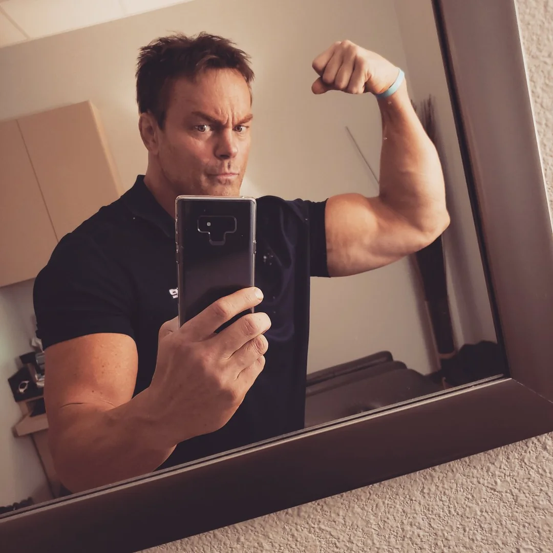 A man takes a selfie in a mirror while flexing his right arm and making a serious face. He is wearing a dark short-sleeve shirt and holding his phone in his left hand. The background shows a room with cabinets and a couch.
