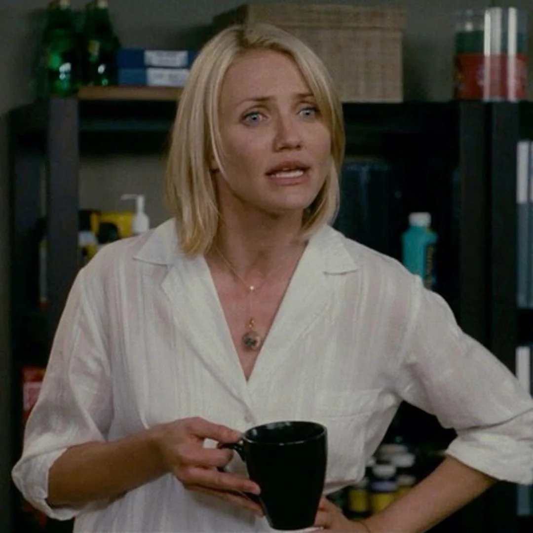 A woman with blonde hair wearing a white shirt holds a black mug and appears to be talking, standing in front of shelves with various bottles and containers.
