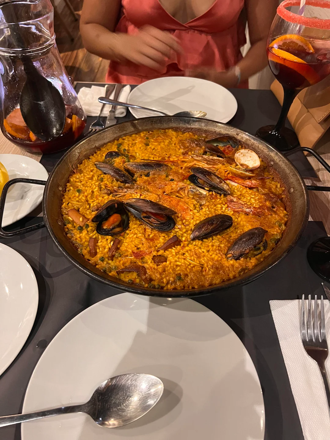 A pan of seafood paella with mussels, rice, and shrimp sits on a table set with plates, cutlery, and sangria. A person in a pink dress is seated in the background.