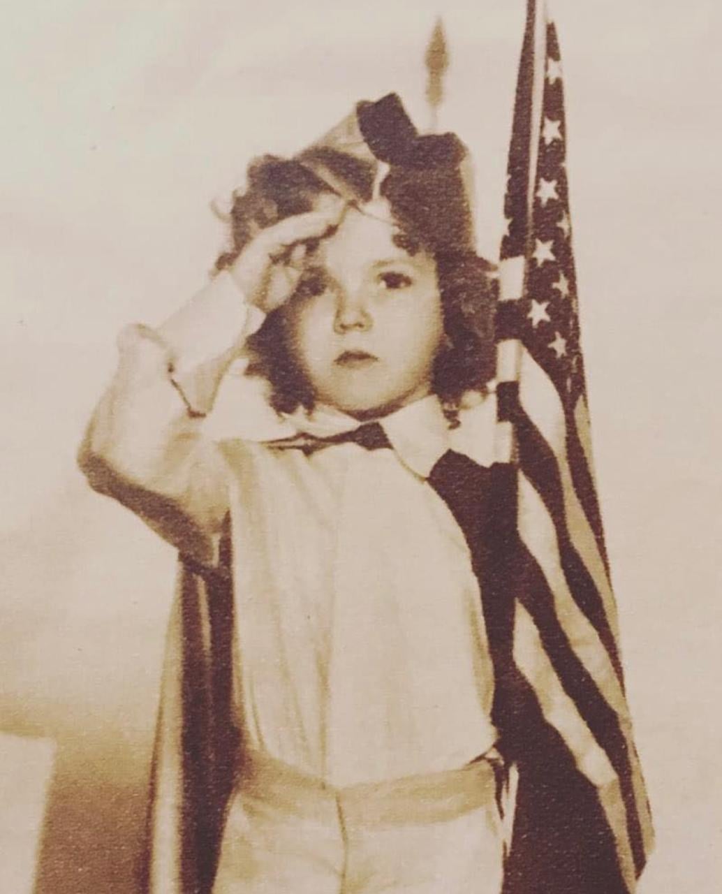 A young child in vintage clothing salutes while holding an American flag. The image is sepia-toned, giving it an old-fashioned, historical appearance.