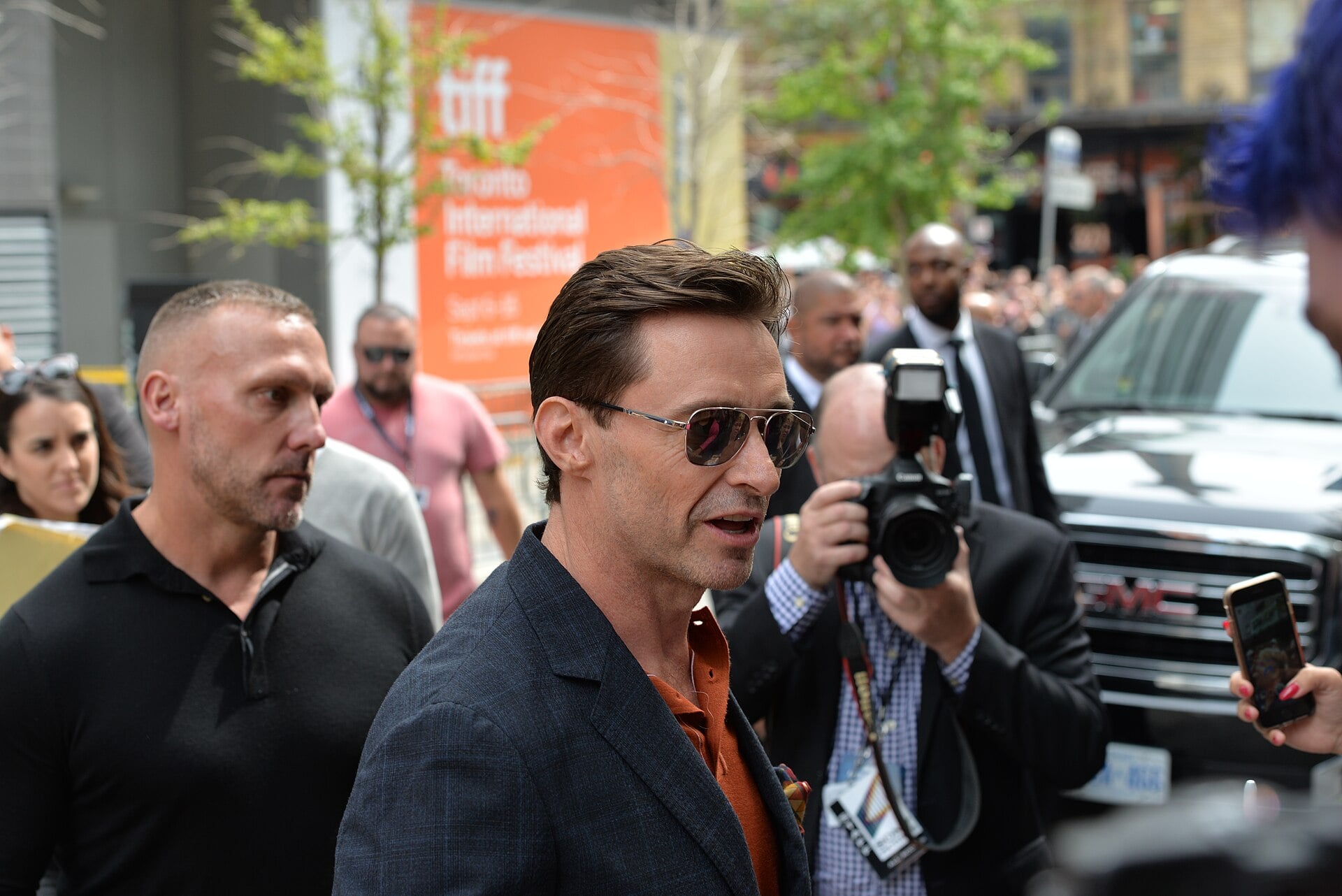 A man in sunglasses and a blazer walks outside surrounded by people, including a photographer taking his picture. In the background, there is a large orange Toronto International Film Festival sign.