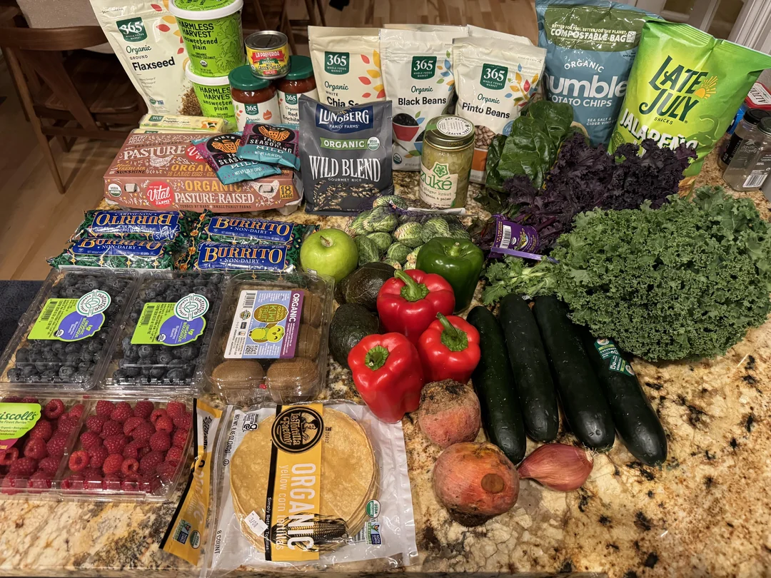A variety of groceries on a kitchen counter, including fresh vegetables, fruits, tortillas, chips, beans, berries, flaxseed, and packaged foods. Items include kale, zucchini, brussels sprouts, peppers, apples, and pomegranate.