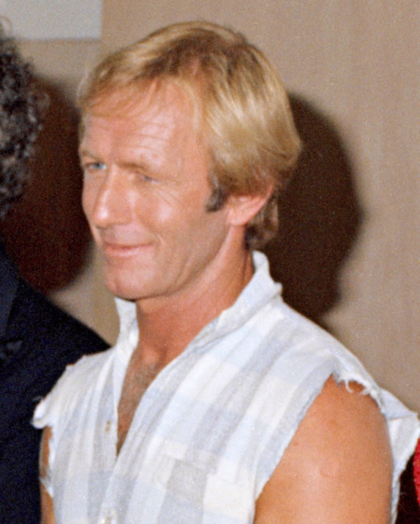 A man with short blond hair, wearing a sleeveless, light plaid shirt, is smiling slightly while standing indoors against a neutral background.