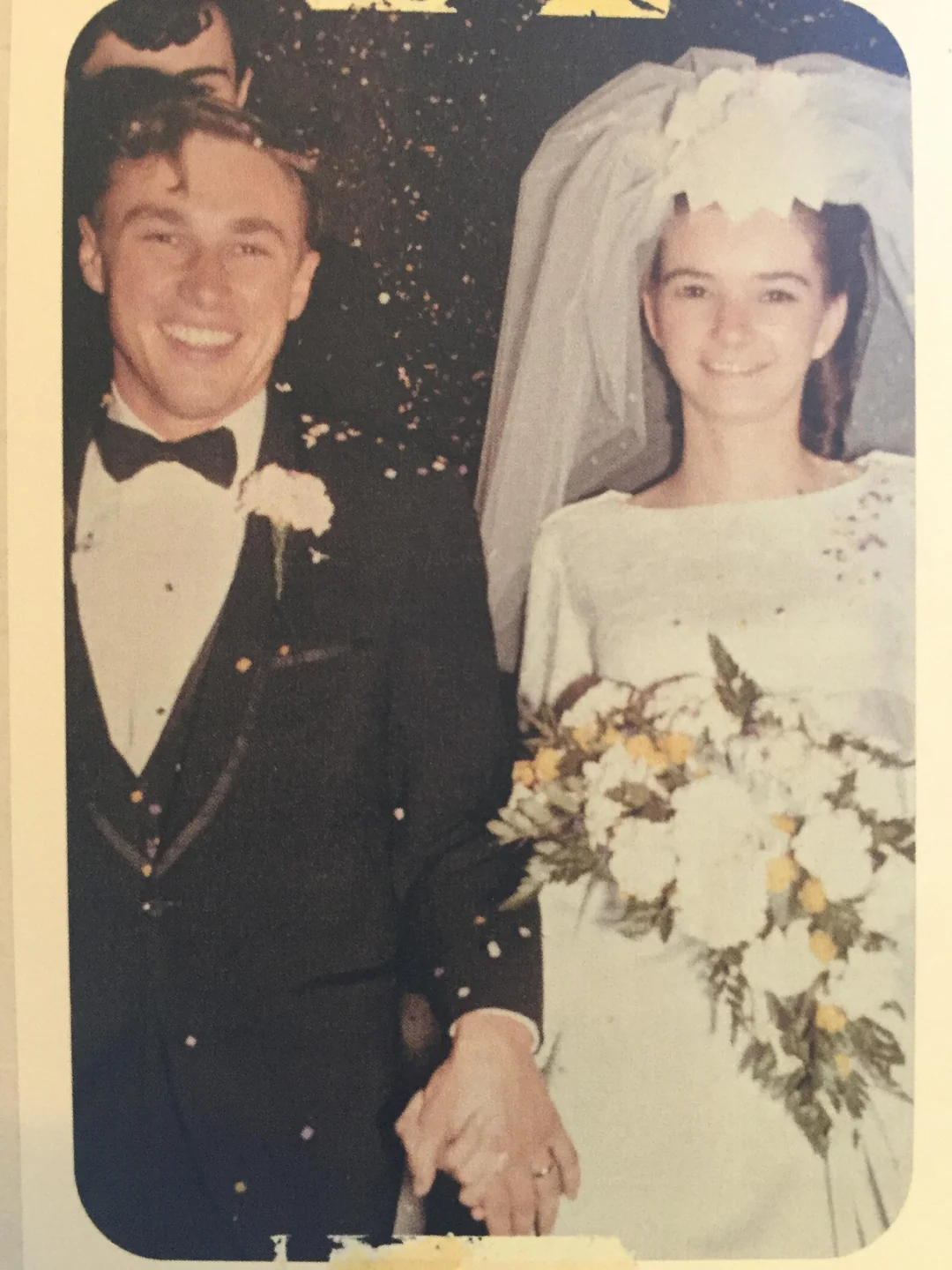 A smiling bride and groom walk hand in hand. The groom wears a black tuxedo, and the bride wears a white dress with a veil and holds a bouquet of white flowers. Confetti floats in the air around them.