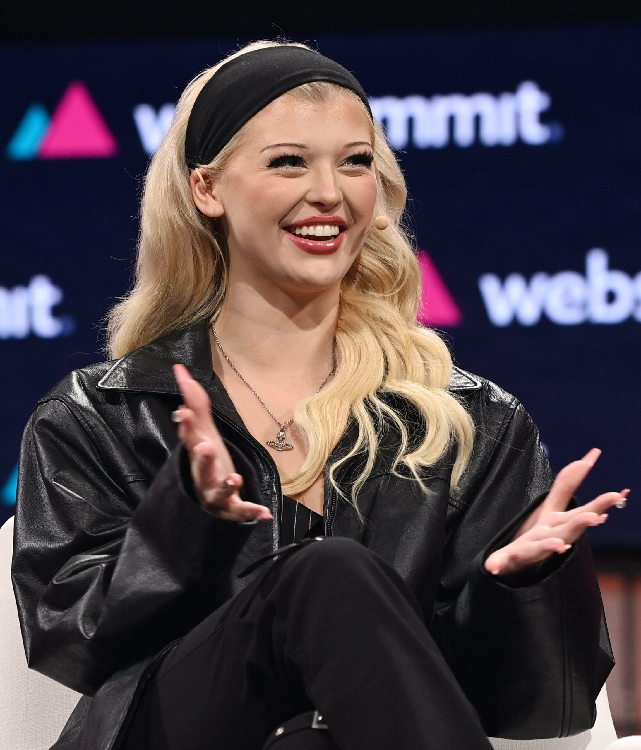 A smiling woman with long blonde hair, wearing a black headband and black leather jacket, gestures with her hands while seated on stage at an event with "web summit" visible in the background.