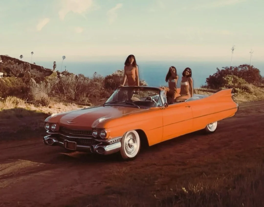 Three women in swimsuits sit in and on top of a vintage orange convertible parked on a dirt road, with coastal scenery and the ocean in the background under a partly cloudy sky.