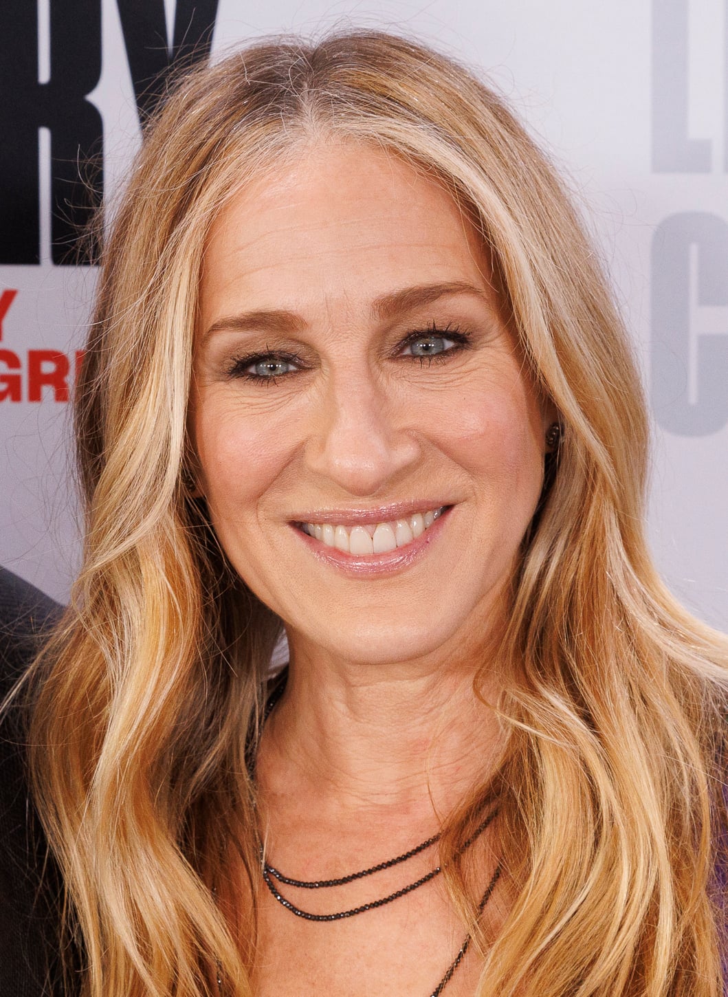 A woman with long, wavy blonde hair smiles at the camera. She is wearing a black top and layered necklaces, and has natural makeup with defined brows. The background is blurred with partial text visible.