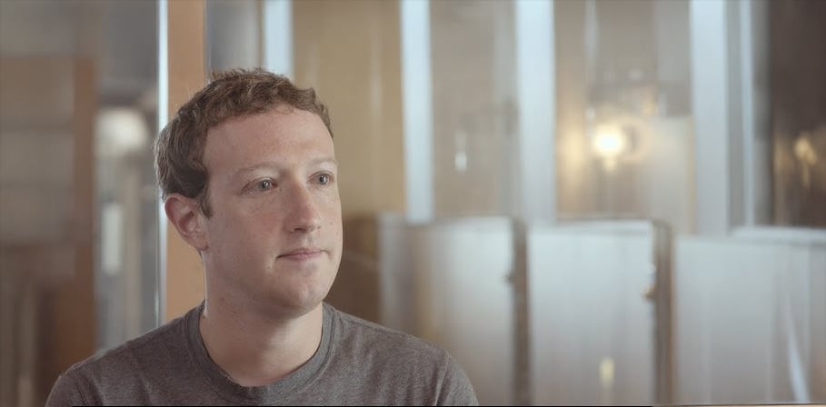 A person with short brown hair and a gray t-shirt sits indoors, looking slightly to the side. The background is softly lit with glass panels and blurred lights.