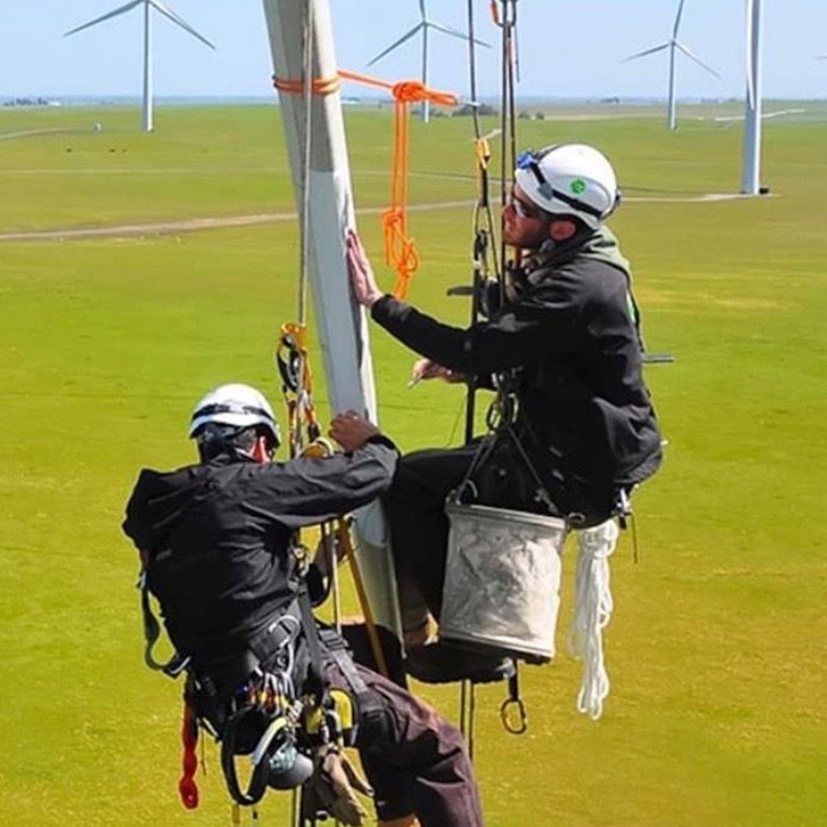 Two workers wearing safety harnesses and helmets perform maintenance on a wind turbine high above a grassy field with several wind turbines in the background.