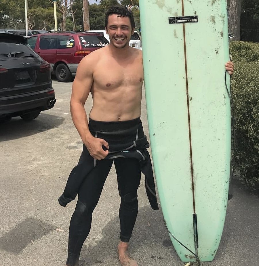 A smiling man in a wetsuit stands barefoot in a parking lot, holding a light green surfboard upright. Cars and trees are visible in the background.