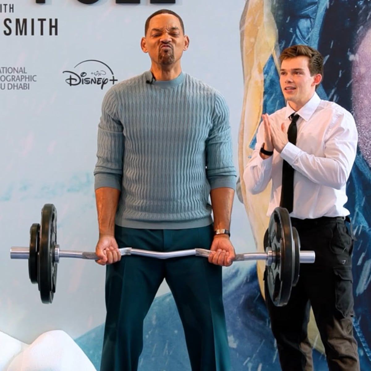A man in a blue sweater lifts a barbell with a strained expression while another man in a white shirt and tie stands beside him, clapping and smiling, in front of a promotional backdrop.