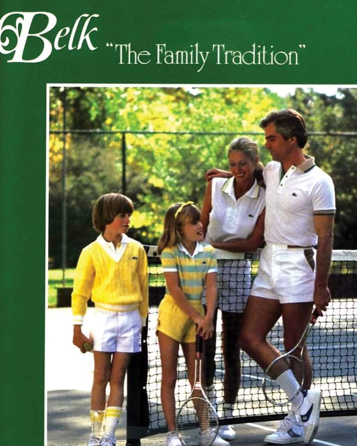 A family of four, dressed in vintage tennis outfits, stands on an outdoor tennis court near the net, smiling and talking. The image is part of a "Belk" advertisement titled "The Family Tradition.