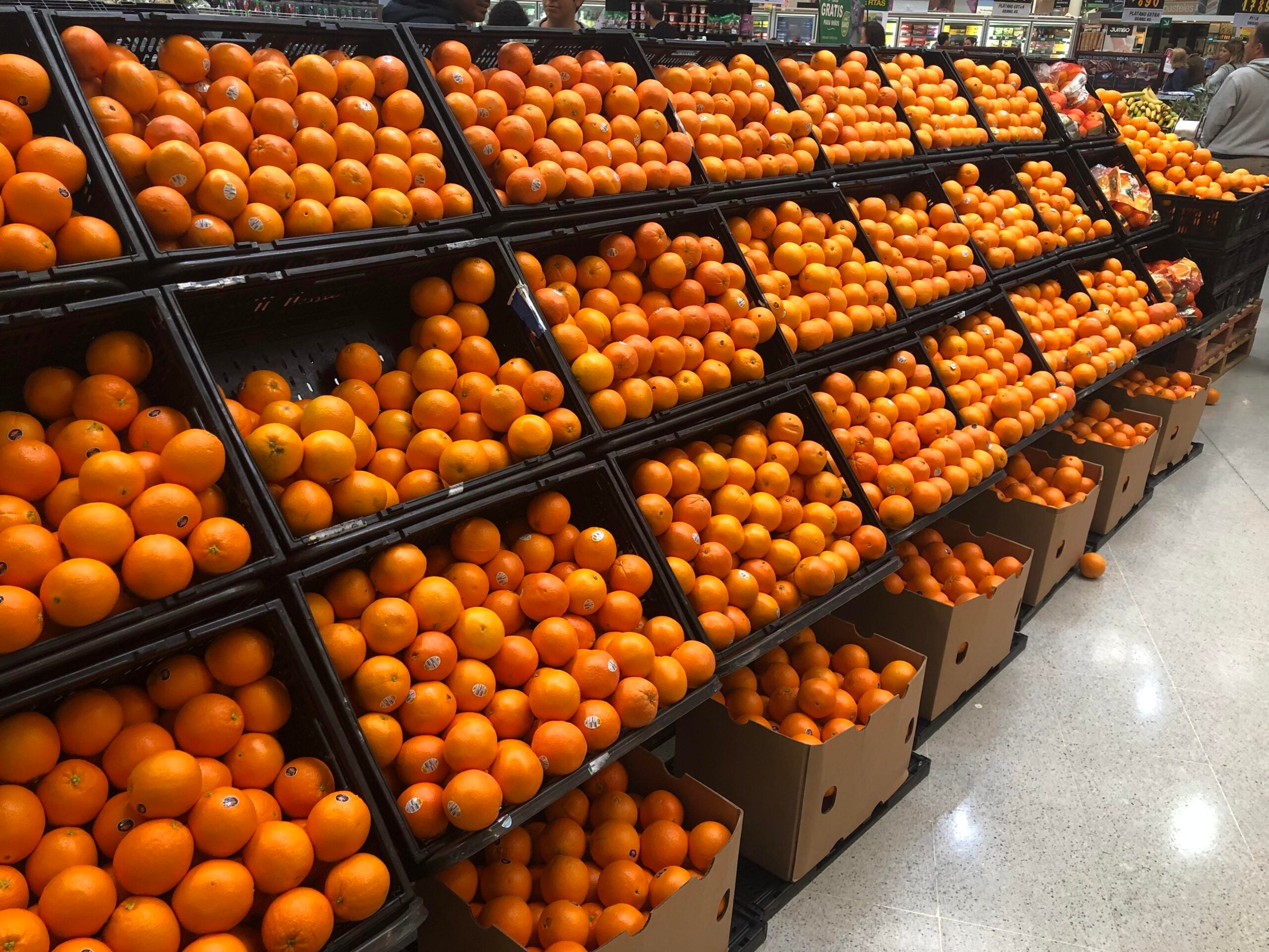Stacks of oranges fill black display shelves and boxes at a grocery store produce section. The bright oranges are neatly arranged in rows, and a few shoppers are visible in the background.