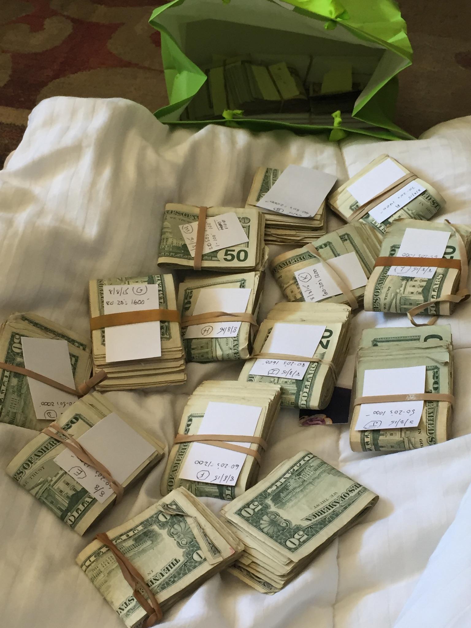 Stacks of U.S. dollar bills, mostly twenties and fifties, bundled with rubber bands and labels, spread out on a bed. A green bag containing more bundles is in the background.