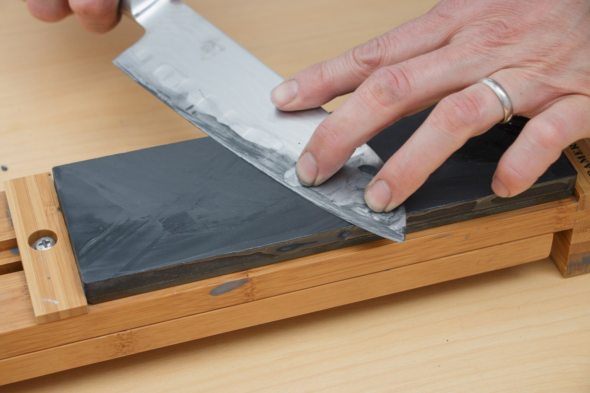 A person sharpens a kitchen knife on a whetstone, holding the blade at an angle with one hand while sliding it across the stone. The setup is on a wooden surface.