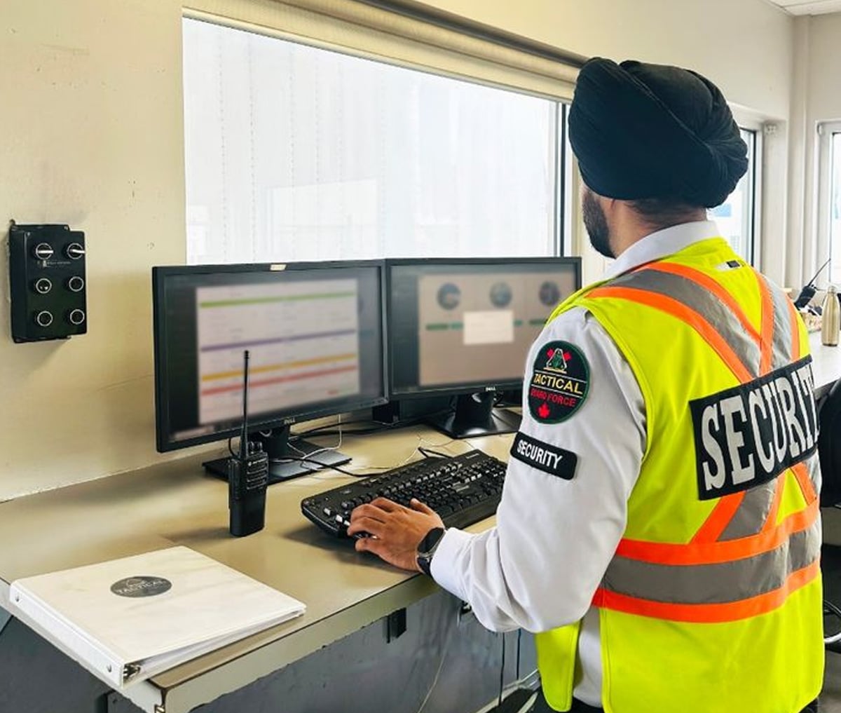 A security guard wearing a high-visibility vest and turban monitors information on two computer screens in an office. A radio and binder are on the desk nearby.