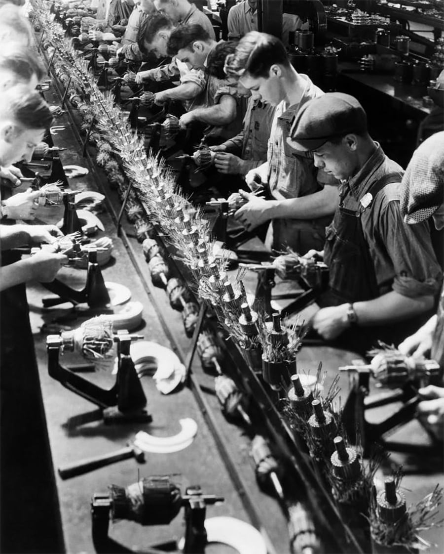Workers assembling machinery components in a crowded factory, standing side by side at long workbenches, focused on their tasks in an industrial setting. The image is black and white, suggesting a historical time period.