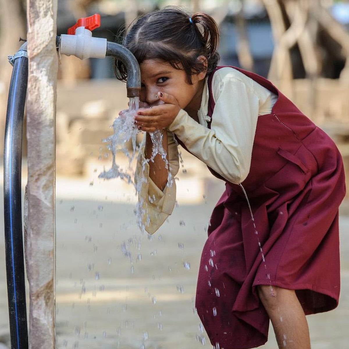 A young girl in a maroon school uniform drinks water from an outdoor tap, cupping her hands to catch the flowing water. She is smiling and bending forward, with water splashing around her.