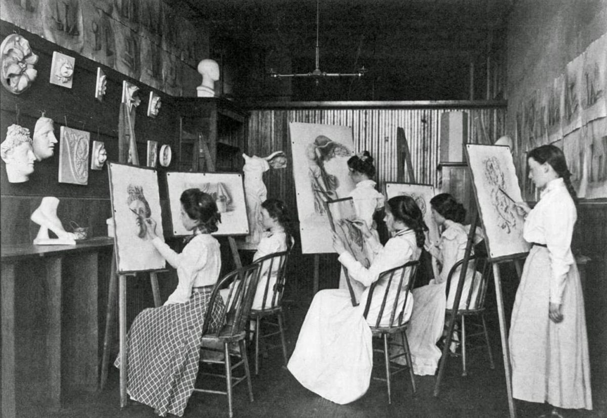 Five women in long skirts and blouses sit in a row at easels drawing classical plaster sculptures in an art studio filled with sketches and busts. One woman stands, observing the others’ work.