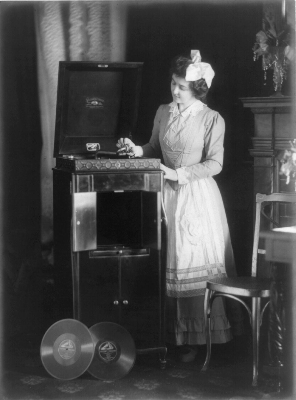 A woman in early 20th-century attire stands beside an open phonograph, preparing to play a record. Two vinyl records are displayed on the floor in front of her, and elegant furnishings are visible in the background.