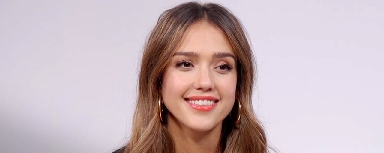 A woman with long, wavy brown hair and hoop earrings smiles in front of a plain, light-colored background.