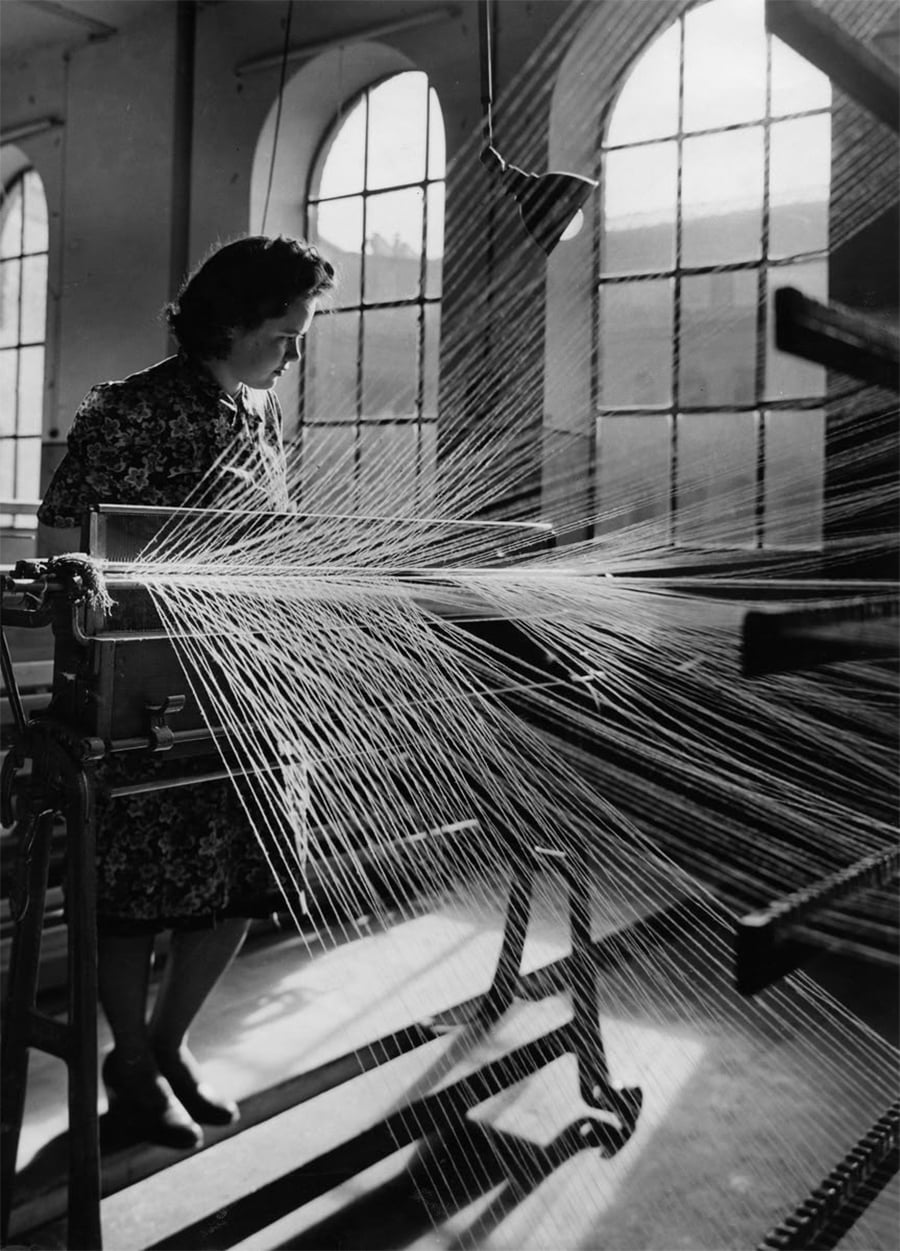 A woman in a patterned dress stands beside a textile machine with numerous threads stretched out, working in a sunlit room with large arched windows.