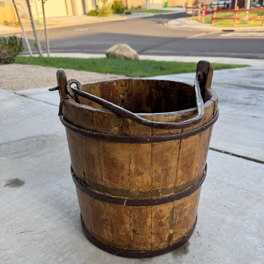 A weathered wooden bucket with metal bands and handle sits on a concrete driveway, with a suburban street and houses visible in the background.